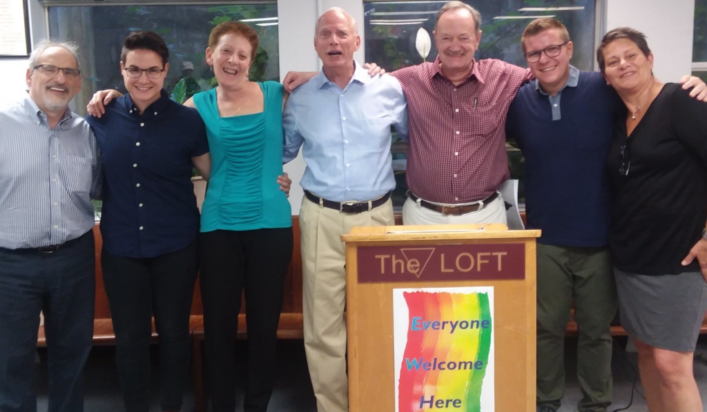 A group shot of participants, teaching artist, and Ed Friedman at the culminating event at The LOFT. The group are standing side by side with their arms around each other in front of a podium.