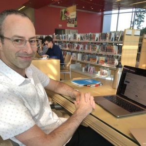 A photo of a man wearing glasses, sitting at a table in a public library. 