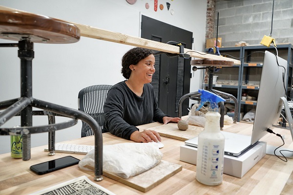 Image of a teaching artist sitting at a desk on a computer. She has clay on the table in front of her. She is smiling at the screen.