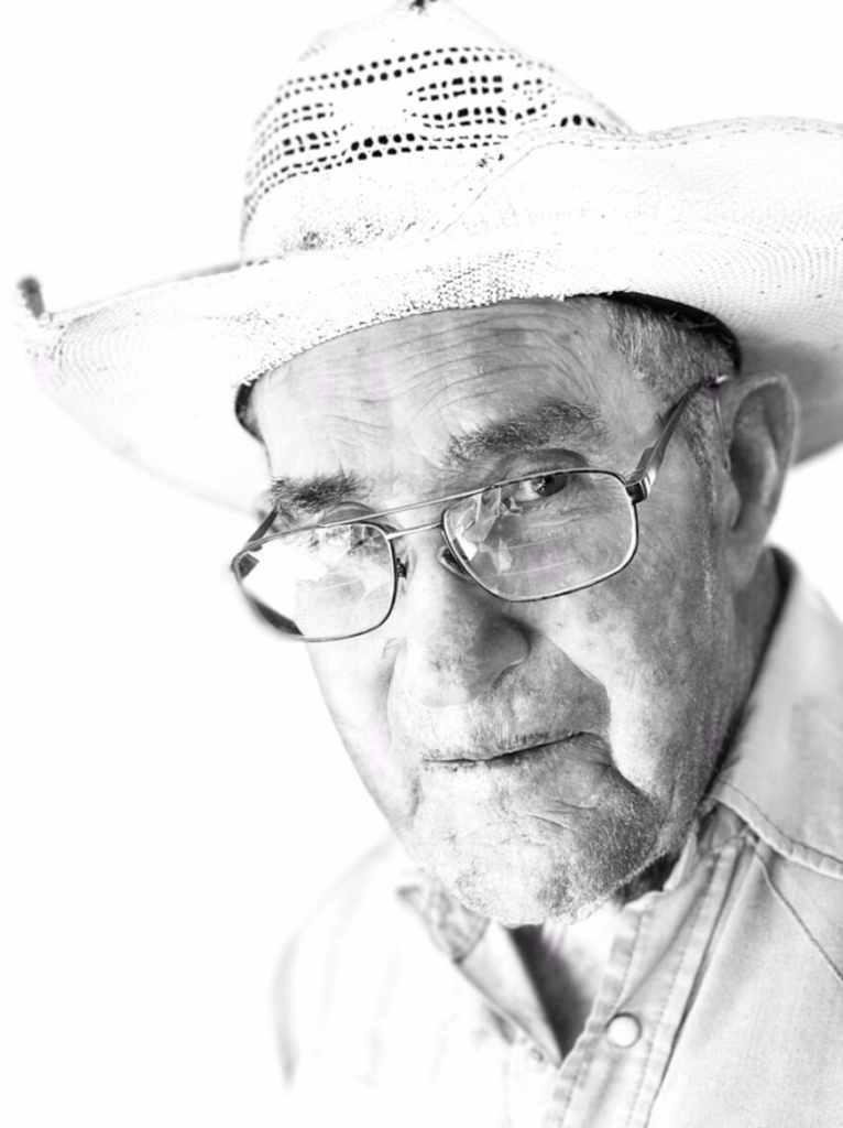 A black and white photo of an older man looking at the camera with glasses and a western hat