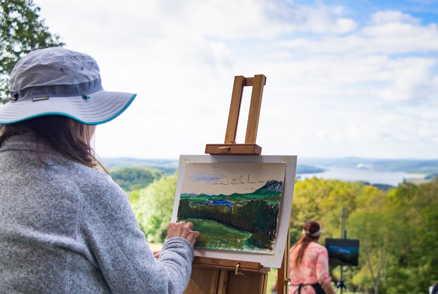 A participant painting on an easel outdoors with a landscape of trees, water, and blue sky in the background.
