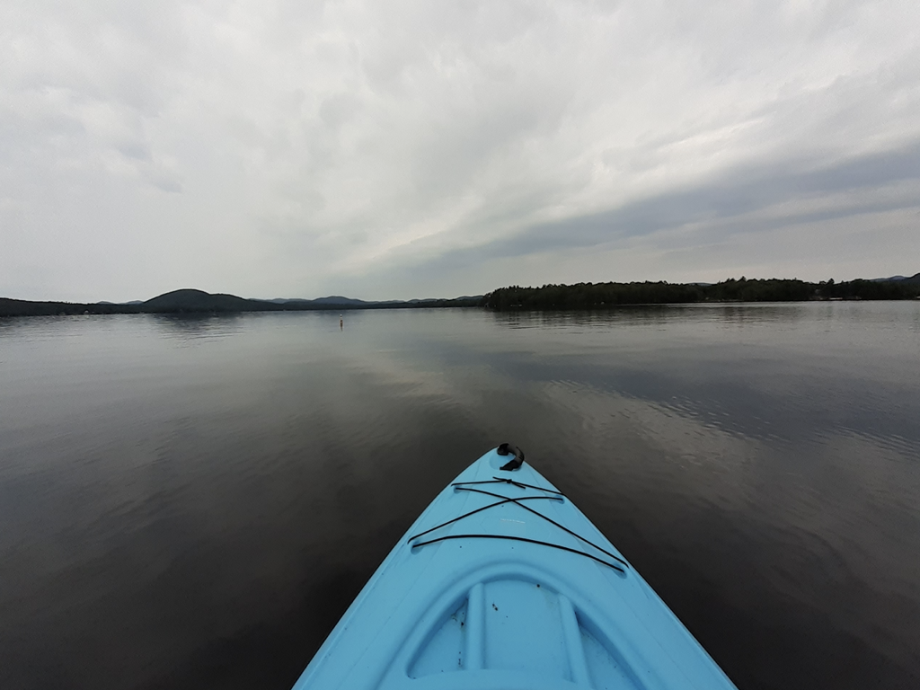 A participant's photograph of a first-person perspective on a blue kayak. The view beyond the kayak is cloudy skies and calm, open water with tree lines in the distance.