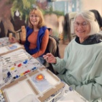 Two older adult participants smile at the camera while painting at a table.