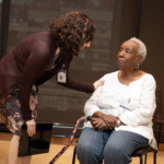 Teaching artist Julie Kline bends down and touches an older adults' arm who is sitting in a chair. Julie is white with dark curly brown hair and is wearing a brown cardigan and floral dress. The older adult woman is black with short white hair and is wearing a white blouse and jeans. She is holding a cane.