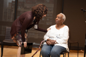 Teaching artist Julie Kline bends down and touches an older adults' arm who is sitting in a chair. Julie is white with dark curly brown hair and is wearing a brown cardigan and floral dress. The older adult woman is black with short white hair and is wearing a white blouse and jeans. She is holding a cane.