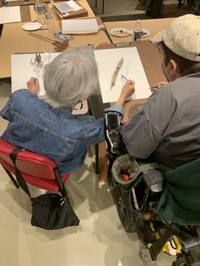 Two older adult students, one sitting in a desk chair and one in a wheel chair, work together on a drawing using drawing boards in their laps